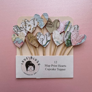 May include: A set of 12 heart-shaped cupcake toppers. Each heart is printed with a vintage map design and attached to a wooden stick. The packaging includes the text "LOVE BLUSH" and "12 Map Print Hearts Cupcake Topper".