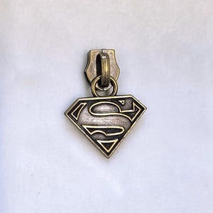 May include: A metal zipper pull featuring the Superman logo. The pull is a diamond shape with the iconic "S" symbol in the center. The metal has a bronze finish, and the pull is attached to a metal loop.