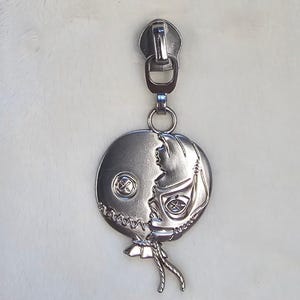 May include: Silver zipper pull with a round charm featuring a stitched-up face with one eye and a button nose.
