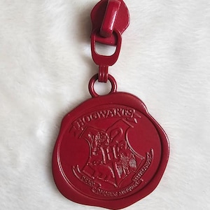May include: Red zipper pull with a Hogwarts seal charm. The seal features the Hogwarts crest with the words "Hogwarts" and "Draco Dormiens Nunquam Titillandus".