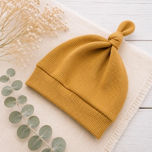 May include: A mustard yellow baby beanie crafted from a textured waffle knit fabric. The hat features a folded brim and a small, knotted top detail. The design is simple and functional, showcasing the grid-like pattern of the fabric clearly.