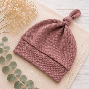 May include: A dusty rose colored baby hat featuring a waffle knit texture and a knotted top detail. The hat has a wide folded brim and is constructed from a soft fabric, showcasing a consistent grid pattern throughout the entire garment design.