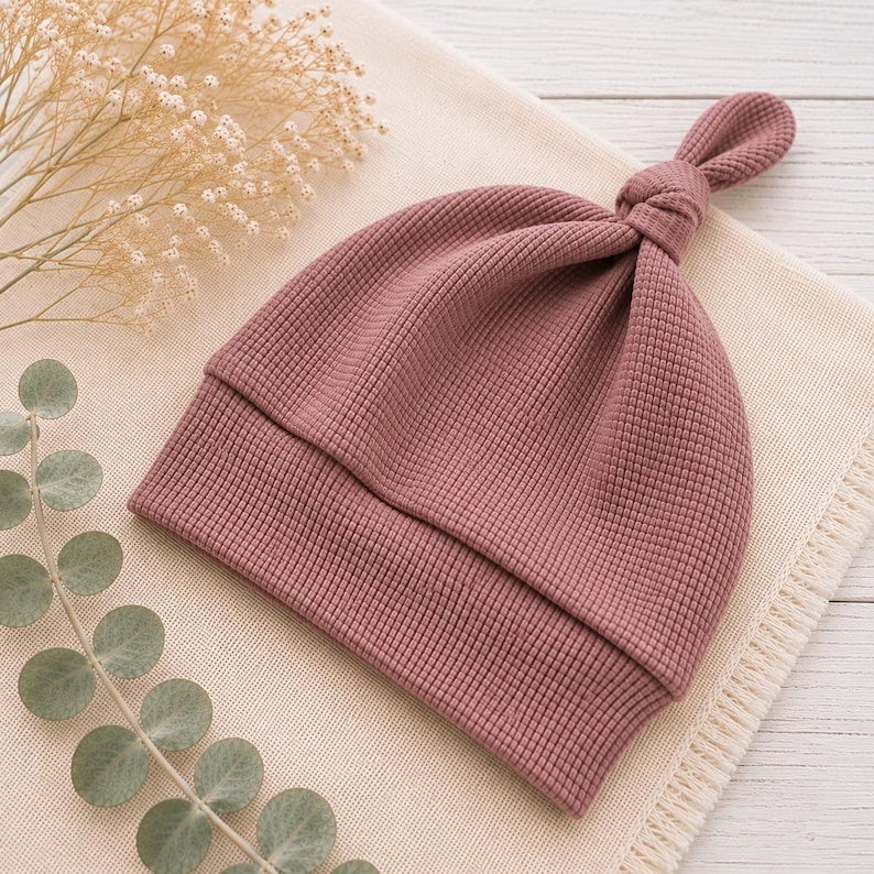May include: A dusty rose colored baby hat featuring a waffle knit texture and a knotted top detail. The hat has a wide folded brim and is constructed from a soft fabric, showcasing a consistent grid pattern throughout the entire garment design.