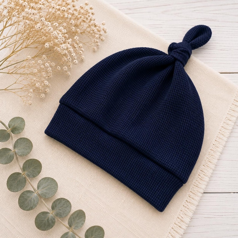 May include: A dark navy blue beanie hat crafted from a textured waffle-knit fabric. The hat features a folded brim and a small, knotted top detail, providing a simple and functional design for headwear that is both soft and durable in construction.