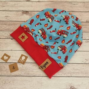 May include: A blue and red beanie with a fire truck pattern. The beanie has a red fold-over cuff and a brown star-shaped tag. The beanie also has a brown tag with the name "Fynn" on it.
