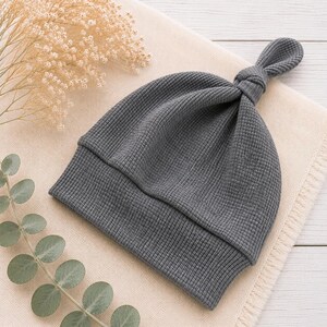 May include: A dark gray beanie hat crafted from a textured waffle-knit fabric. The hat features a folded brim and a knotted top detail, creating a distinct silhouette. The material has a consistent grid-like pattern throughout the entire design.