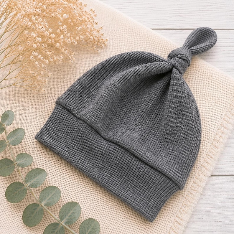 May include: A dark gray beanie hat crafted from a textured waffle-knit fabric. The hat features a folded brim and a knotted top detail, creating a distinct silhouette. The material has a consistent grid-like pattern throughout the entire design.