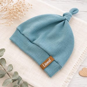 May include: A blue waffle-knit beanie with a knotted top and a brown rectangular faux leather tag featuring the name Lionel in white script. The hat has a folded brim and a textured fabric construction, showcasing a simple and functional design.