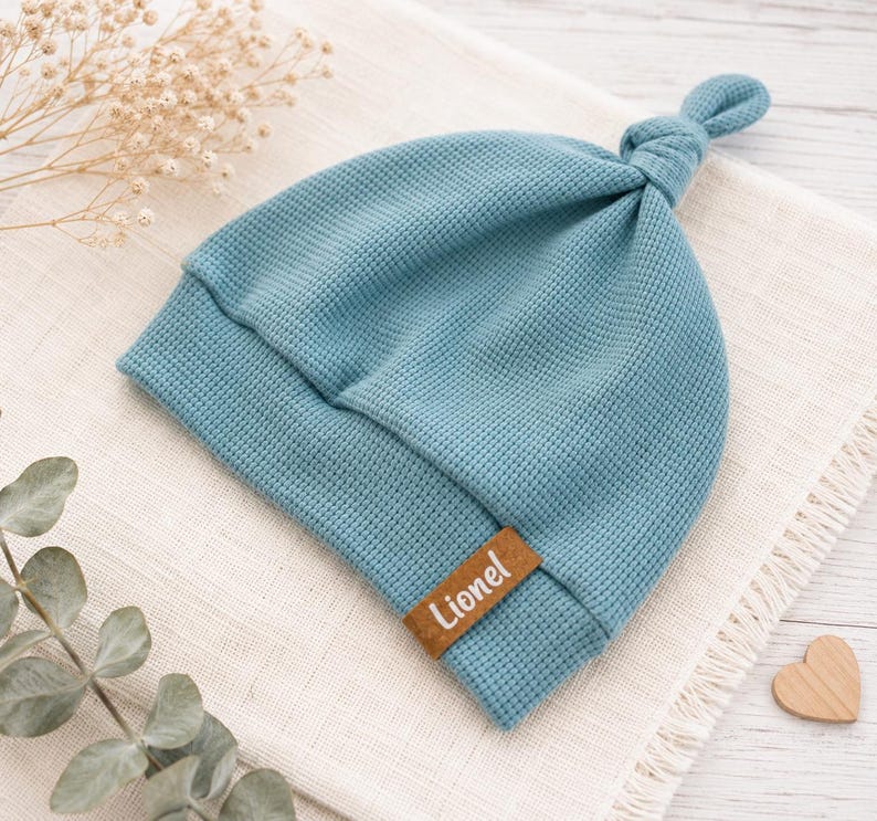 May include: A blue waffle-knit beanie with a knotted top and a brown rectangular faux leather tag featuring the name Lionel in white script. The hat has a folded brim and a textured fabric construction, showcasing a simple and functional design.