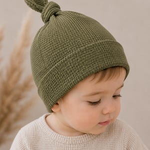 May include: An olive green waffle-knit beanie with a knotted top detail and a folded brim. The hat features a textured grid pattern throughout the fabric, providing a structured look while maintaining a soft appearance for the head covering.