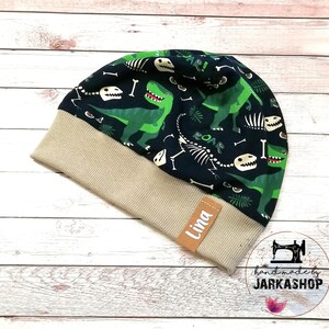 May include: A beige and blue knit beanie with a dinosaur print. The beanie has a brown leather tag with the name "Lina" on it.