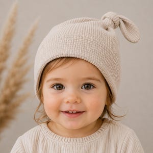 May include: A beige waffle-knit beanie with a knotted top detail. The hat features a textured grid pattern and a folded brim, crafted from a soft fabric. The design includes a small knot at the crown, providing a simple and functional head covering.