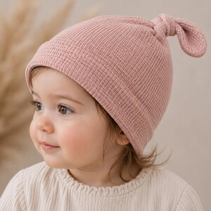 May include: A dusty pink waffle-knit beanie featuring a folded brim and a knotted top detail. The hat is constructed from a textured fabric with a grid-like pattern, providing a soft appearance. The design includes a simple knot at the crown.