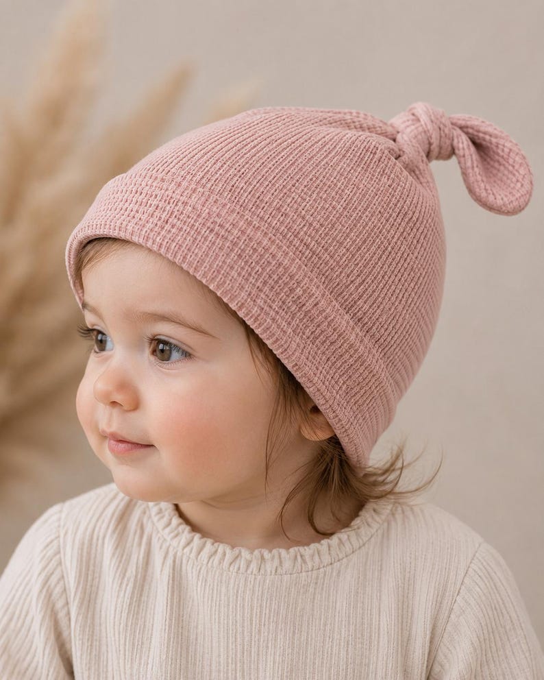 May include: A dusty pink waffle-knit beanie featuring a folded brim and a knotted top detail. The hat is constructed from a textured fabric with a grid-like pattern, providing a soft appearance. The design includes a simple knot at the crown.