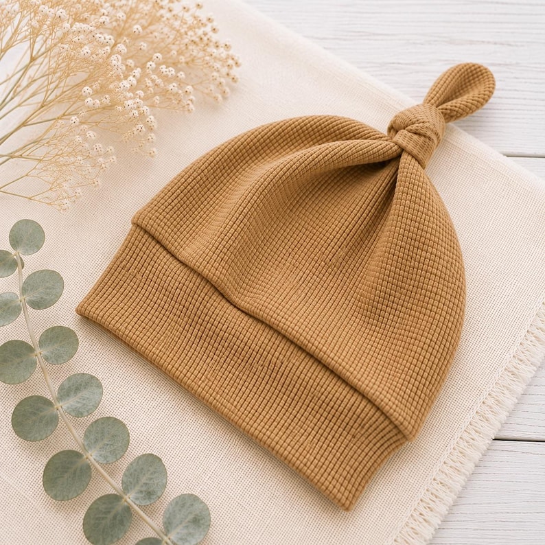 May include: A mustard yellow baby hat made of a textured waffle knit fabric. The hat features a folded brim and a knotted top detail. The design is simple and functional, showcasing the grid-like pattern of the fabric and the solid color.