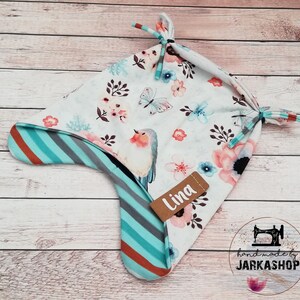 May include: A baby bonnet with a floral and bird print. The bonnet features a white background with pink, blue, and coral flowers, butterflies, and a bird. The ear flaps have a striped pattern in teal, gray, and brown. The label says "Lina".