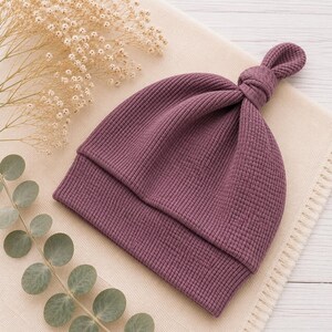 May include: A deep plum-colored beanie hat crafted from a textured waffle-knit fabric. The hat features a folded brim and a decorative knotted top detail, showcasing a consistent grid-like pattern throughout the material of the headwear.