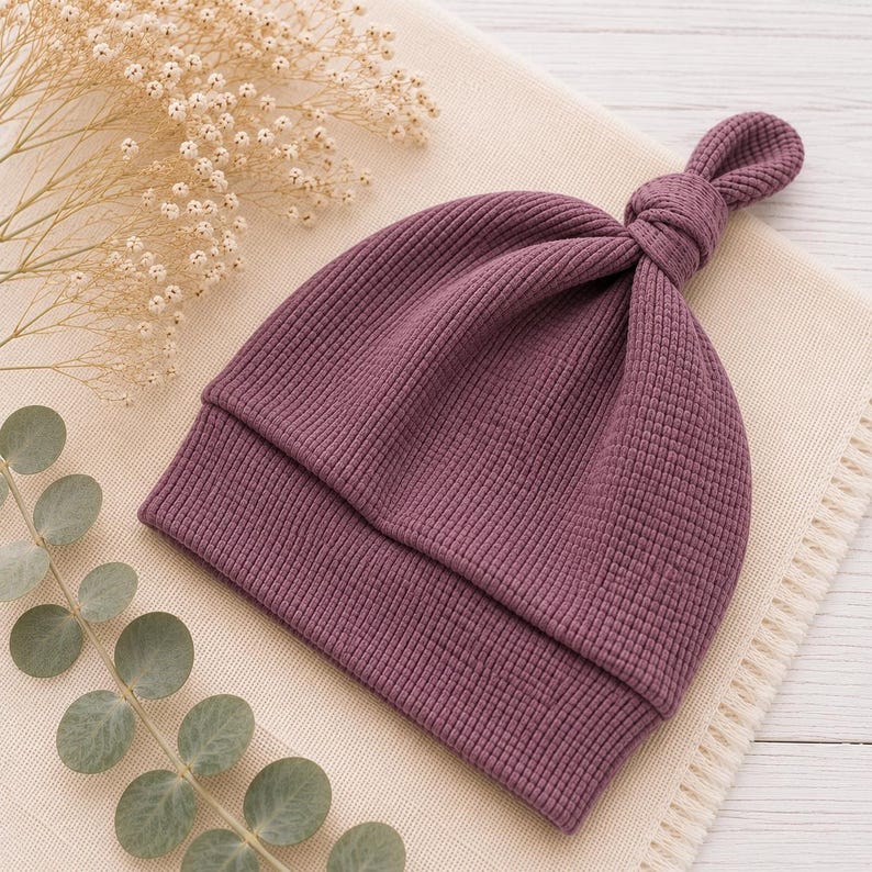 May include: A deep plum-colored beanie hat crafted from a textured waffle-knit fabric. The hat features a folded brim and a decorative knotted top detail, showcasing a consistent grid-like pattern throughout the material of the headwear.