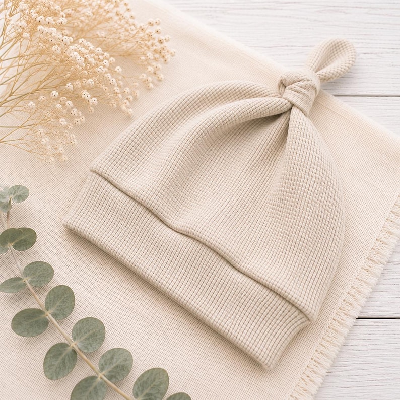 May include: A beige waffle-knit beanie with a knotted top and a wide folded brim. The hat features a textured grid pattern throughout the fabric, creating a soft and uniform appearance. The design is simple, featuring a single knot at the crown.