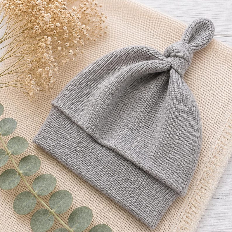 May include: A light gray knit hat featuring a ribbed texture and a knotted top detail. The hat has a folded brim and is constructed from a soft, flexible fabric. The design is simple and functional, showcasing a consistent knit pattern throughout.