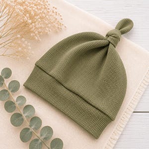 May include: An olive green baby hat featuring a textured waffle knit fabric and a knotted top detail. The beanie has a wide folded brim and a soft, flexible construction, designed with a simple pull-on style for a comfortable and secure fit.