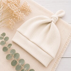 May include: A cream-colored baby hat made of a textured waffle-knit fabric. The hat features a folded brim and a knotted top detail, creating a soft and simple design. The material has a distinct grid-like pattern throughout the entire surface.