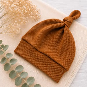 May include: A solid rust-colored beanie made from a textured waffle-knit fabric. The hat features a folded brim and a decorative knot detail at the top center, creating a distinct gathered appearance in the fabric construction.