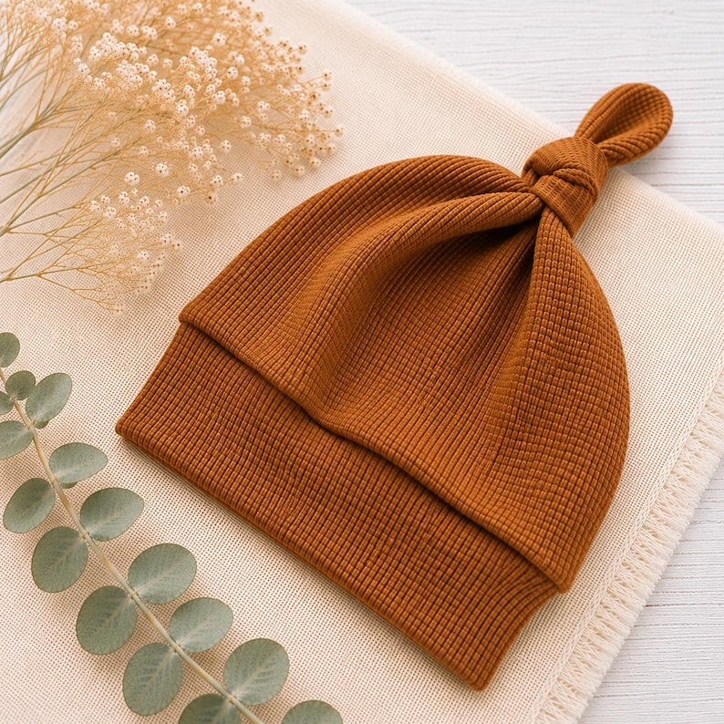 May include: A solid rust-colored beanie made from a textured waffle-knit fabric. The hat features a folded brim and a decorative knot detail at the top center, creating a distinct gathered appearance in the fabric construction.