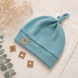 May include: A teal blue waffle-knit beanie with a knotted top and a folded brim. A small square tan leather patch featuring a cutout heart is attached to the front. Three additional small square tan patches with anchor, star, and skull icons.