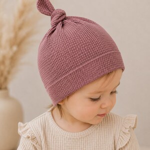May include: A dusty rose waffle-knit beanie with a knotted top detail. The hat features a textured grid pattern and a wide, folded brim that sits snugly around the head. The fabric has a soft, matte appearance with a consistent deep pink hue.