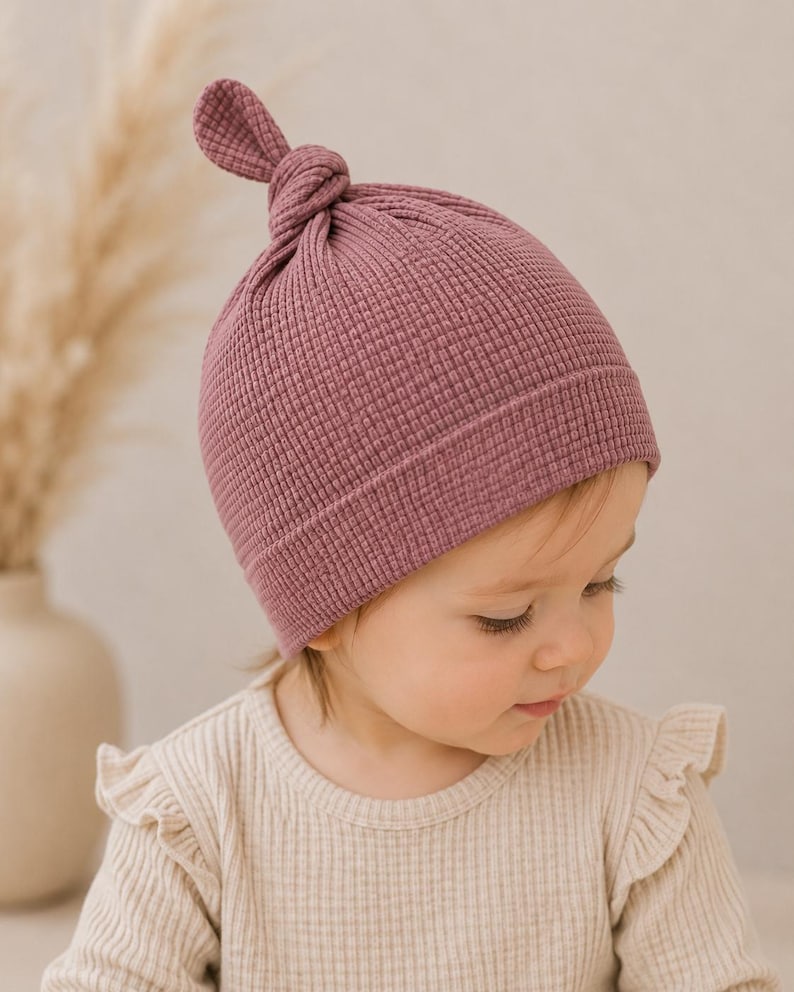 May include: A dusty rose waffle-knit beanie with a knotted top detail. The hat features a textured grid pattern and a wide, folded brim that sits snugly around the head. The fabric has a soft, matte appearance with a consistent deep pink hue.