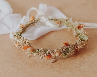 May include: A delicate floral crown made of dried flowers in shades of white, beige, and orange. The crown is lying on a brown surface.