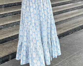 May include: A woman wearing a white tank top and a blue and white tiered skirt with a floral print. She is wearing white sandals and has her hair down.