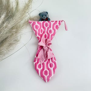 Op de afbeelding: Een roze en witte stropdas met patroon en bijpassende strik, hangend aan een witte muur. Een kleine grijze teddybeer zit bovenop de stropdas. Een rood-wit gestreepte stof bevindt zich boven de stropdas.