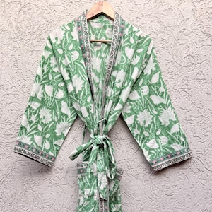 May include: A green and white floral patterned robe hanging on a wooden hanger. The robe has a tie closure and decorative trim along the edges. The floral pattern is a mix of white flowers and green leaves.