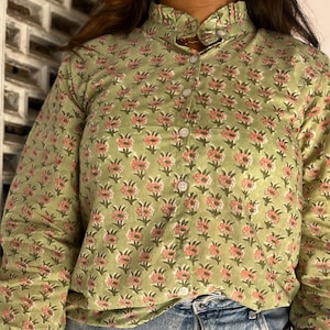 Puede incluir: Una camisa de manga larga con botones en color verde salvia claro con un patrón repetitivo de pequeñas flores rosas y hojas verdes. La camisa tiene un cuello alto con volantes y puños con volantes, cada uno con un botón blanco.