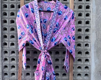May include: A pink floral print robe with blue flower accents and a blue patterned trim. The robe is hanging on a wooden hanger. The robe is tied at the waist with a matching fabric belt.