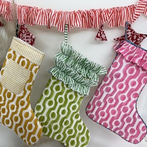 Könnte beinhalten: Drei Weihnachtsstrümpfe in Senfgelb, Grün und Pink, jeweils mit einem einzigartigen geometrischen Muster. Die Strümpfe sind mit Rüschen verziert und hängen unter einer rot-weiß gestreiften Girlande mit sternförmigen Ornamenten.