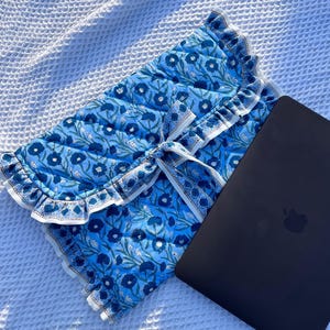 Peut inclure: Une housse d'ordinateur portable bleue à motifs floraux, avec un bord à volants et une fermeture à nouer assortie. Un ordinateur portable noir est posé à côté de la housse sur une surface blanche texturée. Le motif floral présente des fleurs bleu foncé et clair.