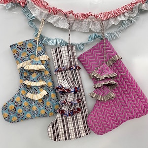 May include: Three decorative Christmas stockings in various patterns and colors. One is blue with yellow floral designs, another is plaid, and the third is pink with a chevron pattern. Each stocking has ruffled trim.