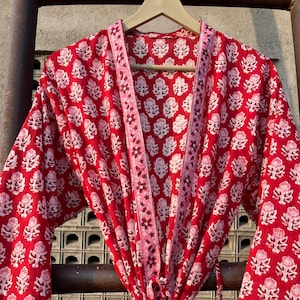 May include: A red cotton robe with a white floral print and a pink trim. The robe has a tie belt and long sleeves.