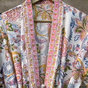 Op de afbeelding: Een witte katoenen badjas met een roze en gele bloemenprint. De badjas heeft een lange, vloeiende silhouet en een strik sluiting in de taille. De stof is zacht en comfortabel, perfect om in huis te ontspannen.