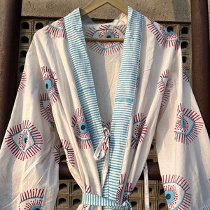 May include: White cotton robe with a blue and white striped trim and a red and blue eye pattern. The robe has a tie belt and two pockets.