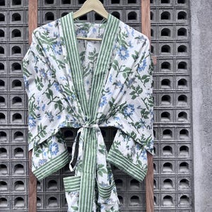 Könnte beinhalten: Ein weißes Bademantel mit Blumenmuster mit blauen Blumen und grünen Blättern. Der Bademantel hat grüne Verzierungen und einen passenden Gürtel. Der Bademantel hängt an einem Holzbügel vor grauem Hintergrund.