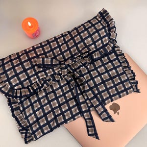 May include: A navy and brown checked laptop sleeve with a frilled edge and bow fastening. The sleeve is on a pink laptop. An orange candle with a lit wick is in the background.