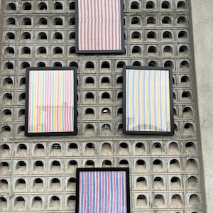 Puede incluir: Cuatro piezas de tela enmarcadas con diferentes patrones de rayas. Los marcos son negros y la tela es colorida. Los marcos están colgados en una pared de concreto con un patrón de cuadrícula.