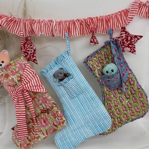 Pode incluir: Três meias de Natal com padrões únicos. Uma é listrada em vermelho e branco, outra em azul e branco, e a terceira é verde com um padrão floral. Cada meia tem um pequeno animal de pelúcia dentro.