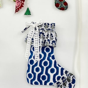 Könnte beinhalten: Ein Weihnachtsstrumpf aus Stoff mit blau-weißem geometrischem Muster, einem gerüschten Umschlag und einer Zehenpartie mit blau-weißem Blumenmuster. Ein weißes Spitzenband ist oben am Umschlag zu einer Schleife gebunden, ergänzt durch kleine Ornamente.