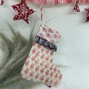 Könnte beinhalten: Ein Weihnachtsstrumpf mit einem rosa-goldenen Blumenmuster, verziert mit einer weißen Manschette und einem dunkelroten Besatz. Er hängt unter einer rot-weiß gestreiften Girlande und sternförmigen Dekorationen.