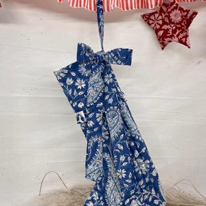 Könnte beinhalten: Ein blauer Weihnachtsstrumpf mit weißem floralem Paisley-Muster, gebunden mit einer Schleife. Ein rot-weiß gestreifter Stoff und ein sternförmiger roter Schmuck hängen darüber. Der Strumpf ist etwa 30 cm lang.